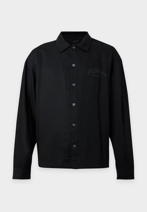 Jeune homme portant une chemise noire à boutons, un pantalon gris et des bottes noires, tenant un sac de voyage en cuir noir par ses poignées.
