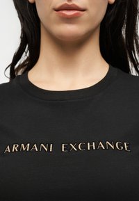 Gros plan d'une personne portant un t-shirt noir avec « ARMANI EXCHANGE » brodé en lettres dorées sur la poitrine.