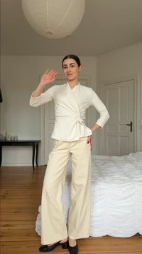Frau in beiger Wickelbluse und weit geschnittenen Hosen winkt, steht in einem Schlafzimmer mit Holzboden und weißer Bettwäsche.
