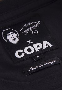 Sorte stofmærke med en hvid silhuet af en mand, signatur og teksten "COPA," med et mindre mærke der siger "Made in Europe."