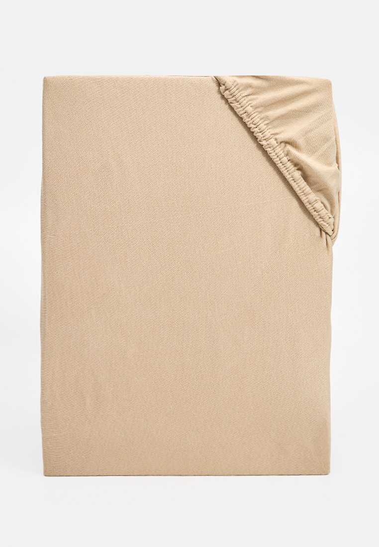 Sábana ajustable beige con bordes elásticos, doblada para mostrar la esquina, sobre un fondo blanco.