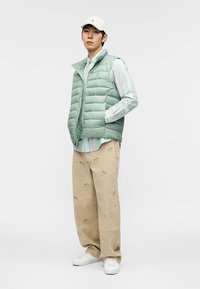 Mężczyzna w białej czapce, miętowej kamizelce puchowej, w paskowaną koszulę, beżowych spodniach z małymi nadrukami zwierząt oraz białych trampkach, stojący na tle jednolitego tła.