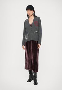Grauer Cardigan mit dekorativen Mustern und Knöpfen, getragen über einem bordeauxfarbenen Samt-Maxi-Kleid. Eine schwarze Mütze und kniehohe schwarze Stiefel runden den Look ab.
