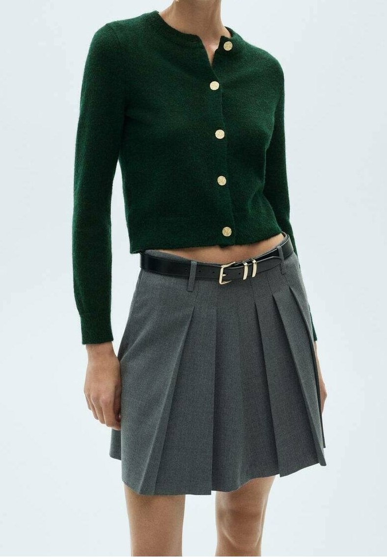 Cardigan en laine vert avec des boutons dorés associé à une jupe plissée grise, avec une ceinture en cuir noir à boucle dorée.
