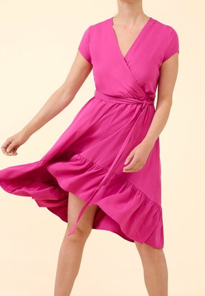 Nainen kirkkaanpinkissä kietaisumekossa, jossa on lyhyet hihat ja röyhelöhelma, seisoo hame hieman kohotettuna ikään kuin liikkeessä pysäytettynä.