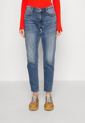 Person iført mid-rise blå jeans, lys rød ribbet sweater og beige plateau sneakers med hvide snørebånd, står på en grå gulv.