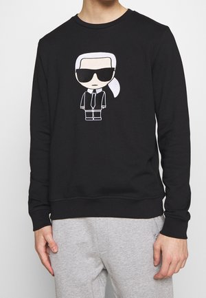 Sudadera negra con un personaje de dibujos animados en el centro, con cabello blanco y gafas de sol. Hecha de un tejido suave y con cuello redondo.