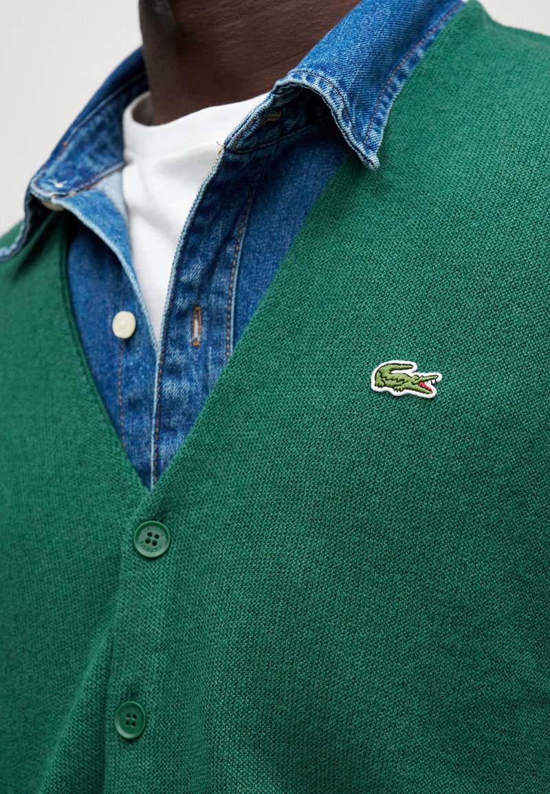 Cárdigan verde de punto con escote en V, con tejido texturizado, cuatro botones y un pequeño logo de cocodrilo bordado en el pecho.