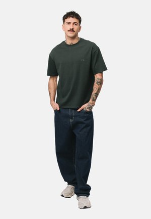 Mężczyzna z tatuażami, ubrany w ciemnozieloną koszulkę, luźne ciemne jeansy i białe trampki, stoi ze rękami w kieszeniach na białym tle.