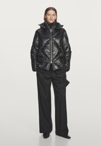 Black puffer jacket with a high collar, featuring a diamond quilted pattern and front zipper, worn with loose black trousers against a white background.
