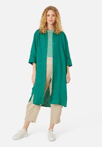 Dlouhá košile z lněné látky v smaragdově zelené barvě s zapínáním na knoflíky, nošená přes pruhované tričko a béžové lněné kalhoty; doplněná bílými teniskami.