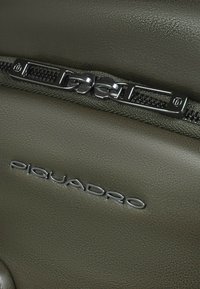 Olijfgroene leren portemonnee met een gestructureerde afwerking, voorzien van een zilverkleurige ritssluiting en het "Piquadro"-logo gegraveerd op het oppervlak.