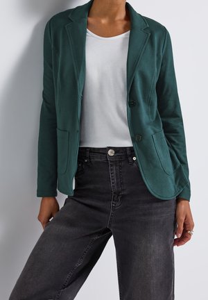 Une personne porte un blazer vert foncé par-dessus un t-shirt blanc et un jean noir taille haute, debout devant un fond clair uni.