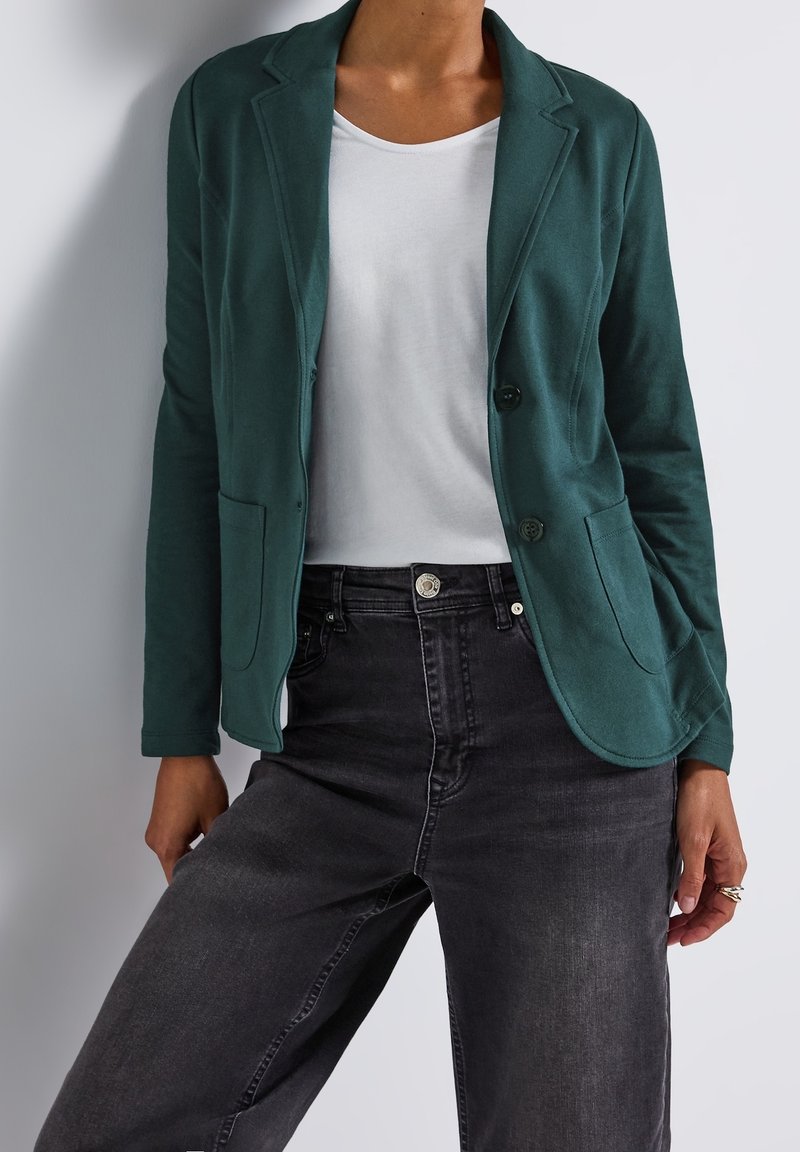 Une personne porte un blazer vert foncé par-dessus un t-shirt blanc et un jean noir taille haute, debout devant un fond clair uni.