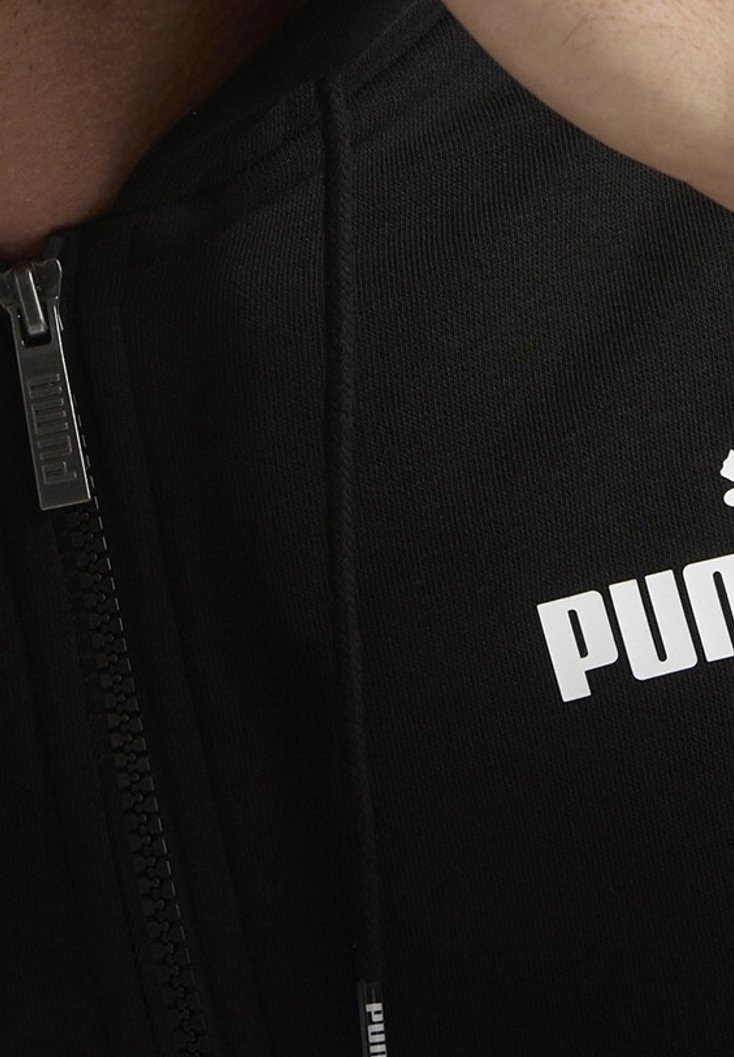 Gros plan sur un sweat à capuche noir Puma montrant la fermeture éclair, le cordon de serrage et le logo partiel blanc Puma sur le tissu.