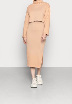 Femme portant un pull court à col roulé beige et une jupe midi assortie avec une fente latérale, associée à des baskets blanches épaisses.