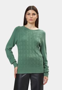 Maglione verde a maglia con motivo a trecce, scollo rotondo e orlo e polsini a coste. Indossato sopra pantaloni di pelle nera.