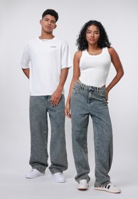 Two individuals stand side by side, wearing oversized jeans and basic tops. The jeans are light blue denim with a loose fit and visible stitching.