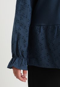 Blusa blu navy con modelli floreali ricamati testurizzati, maniche lunghe arricciate e orlo peplum.