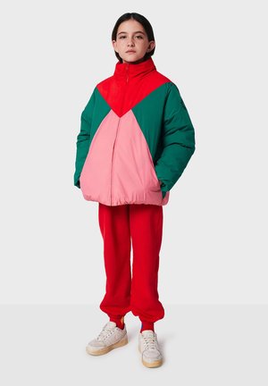 Giubbotto puffer a blocchi di colore con sezioni rosse, verdi e rosa; collo alto; abbinato a pantaloni sportivi rossi e sneakers bianche.