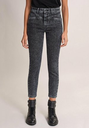 Osoba nosząca czarne skinny jeans z wysokim stanem i efektem acid-wash, połączone z czarnymi sznurowanymi botkami ze sprzączkami.