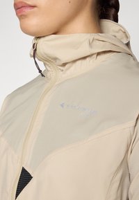 Veste légère beige avec fermeture éclair, dotée d'une capuche, d'un tissu texturé et d'un détail en maille, mettant en valeur une marque réfléchissante.