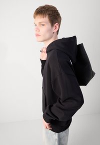 Schwarzer Kapuzenpullover mit lässiger Passform, strukturiertem Stoff und Kängurutasche. Kombiniert mit hellen Denim-Jeans und einer schwarzen Tragetasche.