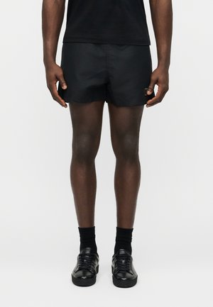 Άνδρας που φοράει μαύρο σορτς, μαύρες κάλτσες και μαύρα δερμάτινα αθλητικά παπούτσια, στέκεται μπροστά σε απλό ανοιχτό φόντο.