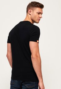 Camiseta negra de manga corta con cuello redondo, hecha de algodón, que presenta una pequeña etiqueta de marca naranja en la manga izquierda. Textura suave.