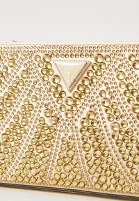Arany tónusú clutch, amely különböző méretű, kiemelkedő sárga rhinestone-okkal rendelkezik, sima felülettel és háromszög alakú logoplakkal.