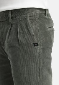 Olive green corduroy pants with vertical ribbed texture, featuring front pleats and a small black logo on the pocket. Classic straight-leg design.