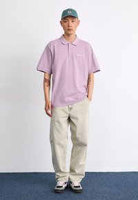Lila Polo-Shirt mit lockerer Passform, graue Jeans und schwarz-pinke Sneakers, ergänzt durch eine türkisfarbene Mütze. Einfaches Design mit minimalem Branding.
