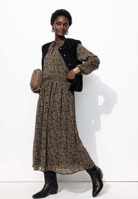 Robe florale à manches longues noire avec des fleurs jaunes, assortie à un gilet noir sans col. Sac à main marron et bottes noires à bout pointu.