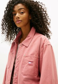 Giacca di jeans rosa con design a colletto, chiusura a bottoni e tasca applicata con etichetta logo rosso e bianco sul petto.