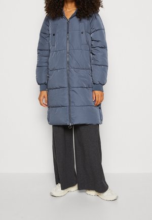 Femme portant une longue doudoune bleue avec fermeture éclair, un pantalon large foncé et des baskets blanches, debout devant un fond blanc uni.