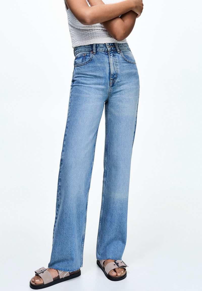 Persona vestida con jeans de tiro alto rectos de color azul claro, un top corto blanco texturizado y sandalias grises tipo deslizantes con hebillas, de pie con los brazos cruzados.