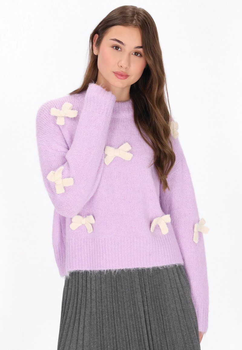 Lavendelfarbener gestrickter Pullover mit langen Ärmeln, versehen mit cremefarbenen Schleifen-Applikationen, geripptem Saum und weicher Textur. Kombiniert mit einem grauen, gefältelten Rock.