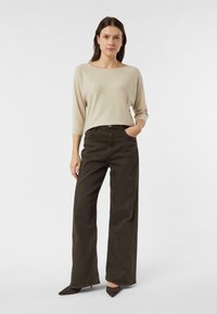 Beżowy, lekko błyszczący sweter z szerokim dekoltem i rękawami 3/4, zestawiony z ciemnozielonymi spodniami o szerokich nogawkach i szpiczastymi ciemnymi butami.