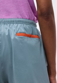 Πίσω όψη ατόμου που φοράει ανοιχτό μπλε παντελόνι με ελαστική μέση, πορτοκαλί τσέπη με περιθώρια και μωβ μπλούζα, με το χέρι να ακουμπάει στο πλάι.