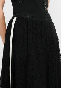 Schwarzer Spitzenrock mit einem strukturierten Blumenmuster, einem breiten elastischen Taillenbund mit "LIU·JO"-Logo und einem kontrastierenden weißen seitlichen Streifen.