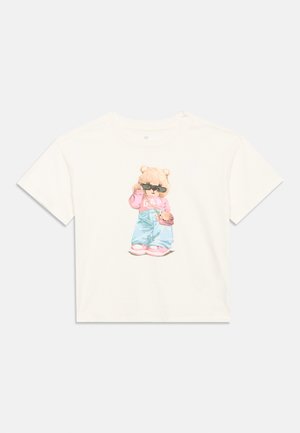 Tricou alb cu mâneci scurte, cu imprimarea unui urs purtând ochelari de soare, hanorac roz, blugi albaștri și pantofi roz în centru.