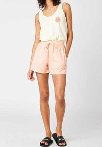 Kobieta ubrana w kremowy top bez rękawów z małym okrągłym graficznym wzorem, jasnoróżowe szorty na gumce w talii oraz czarne klapki Rip Curl.
