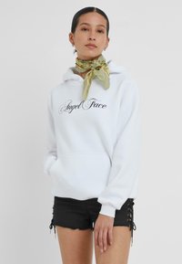 Hoodie blanche avec "Angel Face" imprimé en noir sur le devant, associé à un short noir avec des détails de laçage sur les côtés. Le modèle porte une écharpe verte.