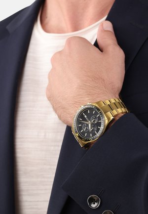 Goldene Uhr mit einem schwarzen Zifferblatt und Chronographenfunktionen, getragen am Handgelenk neben einem marineblauen Blazer. Armbandstil, glänzende Oberfläche und kontrastierende Akzente.
