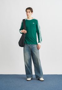 Zelená košeľa s dlhým rukávom s bielymi akcentmi a tromi bielymi pruhmi na rukávoch, v kombinácii so svetlomodrými džínsami s širokými nohavicami a bielymi teniskami.