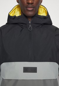 Chaqueta con capucha negra y un panel inferior gris, con un forro interior amarillo, cremallera frontal y parche con logo. Tejido texturizado con capucha ajustable.