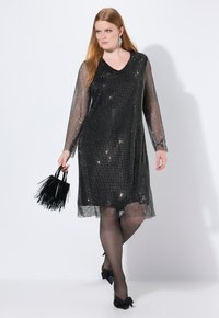 Schwarzes Netzkleid mit langen Ärmeln und V-Ausschnitt, bedeckt mit Pailletten. Kombiniert mit einer schwarzen Handtasche mit Fransen und schwarzen Strumpfhosen sowie Schuhen mit Schleifenakzenten.