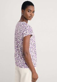 Top de manga corta con estampado floral en tonos morados, detalles en blanco y negro, escote redondo y tejido suave. Corte ajustado, textura ligera.