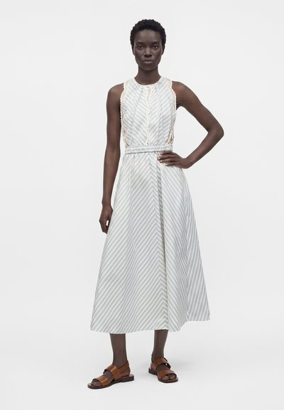 Femme portant une robe midi sans manches ceinturée avec des rayures diagonales gris clair et blanc et des sandales ouvertes marron, debout, une main sur la hanche.