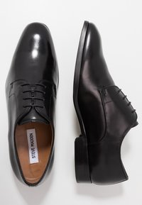 Chaussures de ville en cuir verni noir à lacets, l'une montrée de dessus, l'autre de côté, sur un fond clair et uni.